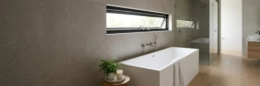 Modern bathroom with a white freestanding bathtub, wooden floor, small round table with plant and candle, and a horizontal window.